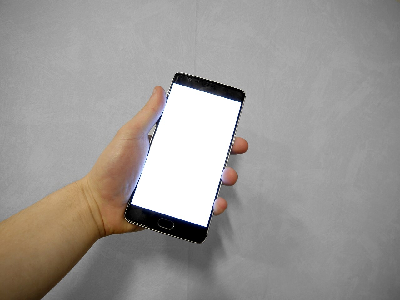 Bàn tay cầm một điện thoại thông minh có màn hình trắng trên nền tường tối.