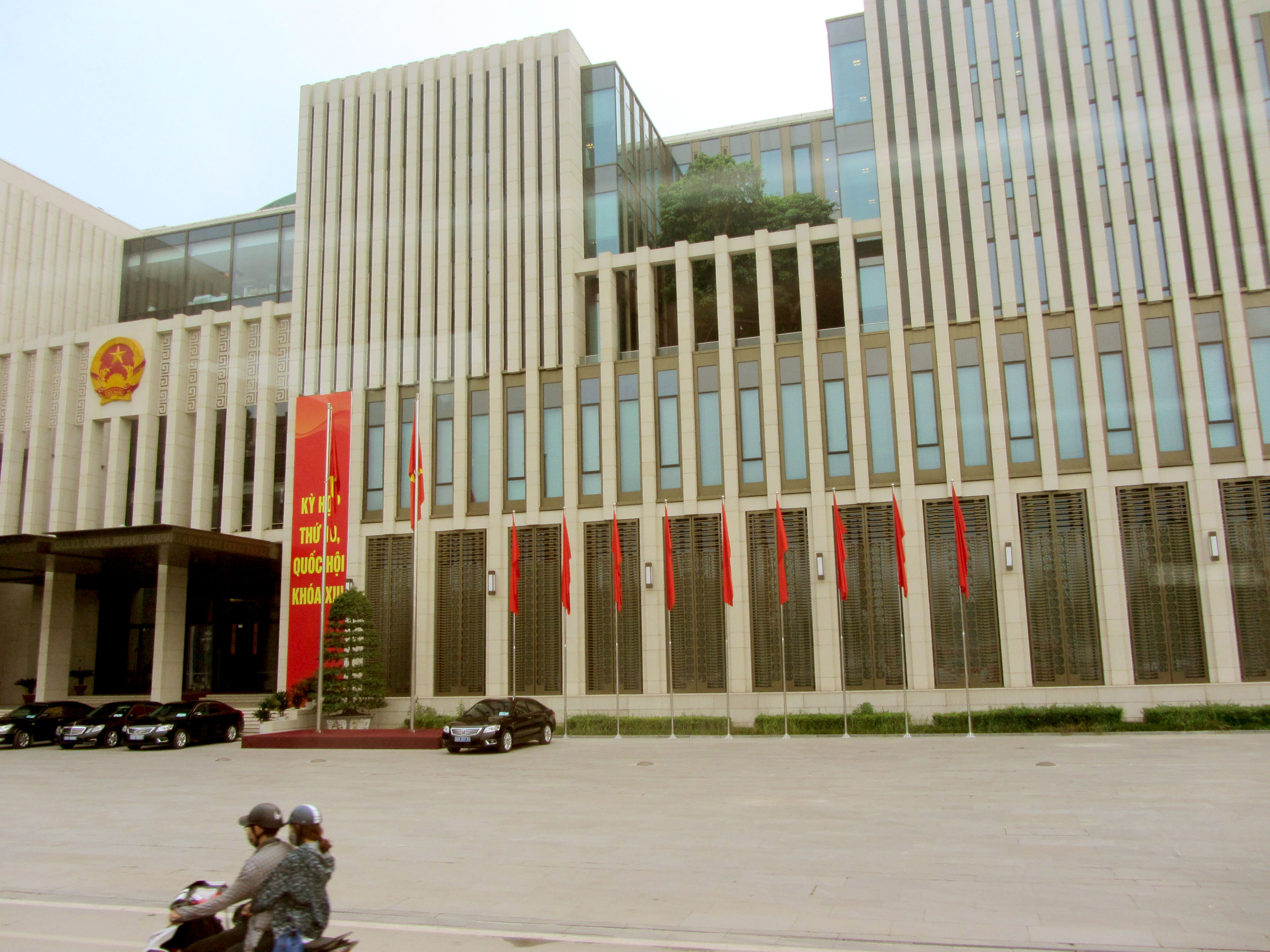 Nhà Quốc hội Việt Nam với quốc huy, nhiều cờ đỏ và người đi xe máy trên đường phía trước.