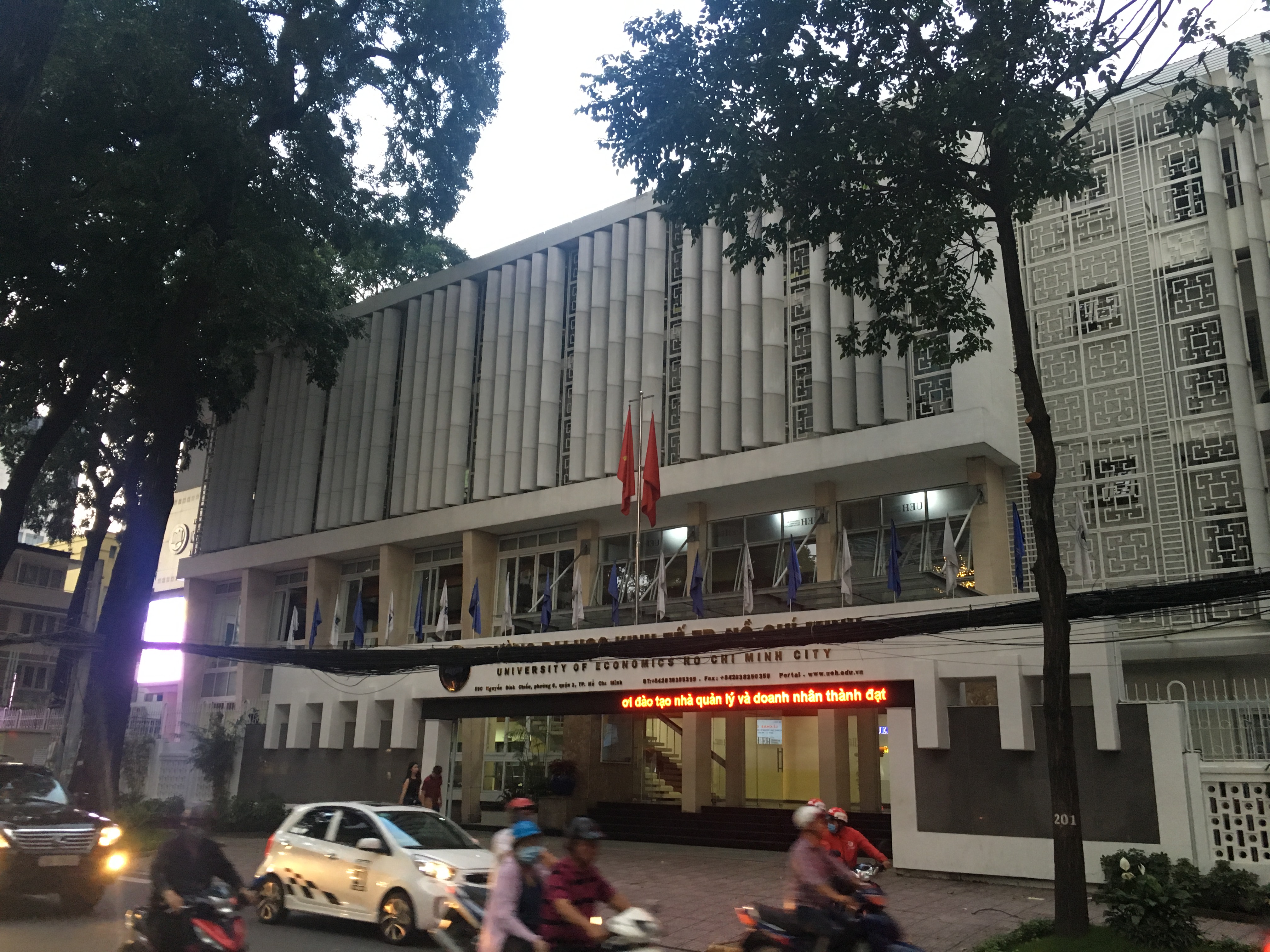 Mặt tiền tòa nhà UEH với cây xanh phía trước, xe máy và ô tô lưu thông trên đường.